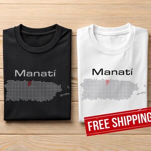 May include: Two t-shirts, one black and one white, each featuring the word "Manatí" above a dotted outline of Puerto Rico. The black shirt has a red dot. The white shirt also has a red dot. The shirts are on a wooden surface.