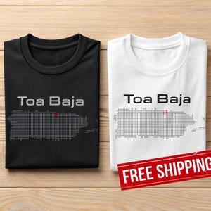 May include: Two folded t-shirts, one black and one white, with the text "Toa Baja" above a dotted outline of Puerto Rico. The black shirt has a grey design, while the white shirt has a grey design. A red "FREE SHIPPING" sticker is visible.