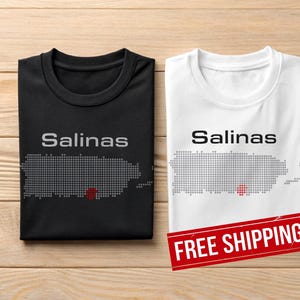 May include: Two t-shirts, one black and one white, with the word "Salinas" printed above a dotted outline of a map. The map has a red dot. The white shirt has a red "FREE SHIPPING" banner.