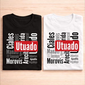 May include: Two t-shirts, one black and one white, each with a design featuring the word "Utuado" in a red rectangle. The design also includes a list of other place names in a stylized font.