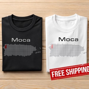 May include: Two t-shirts, one black and one white, each with the word "Moca" above a grey dotted outline of Puerto Rico. A red dot marks a location on the island. The shirts are displayed on a wooden surface.