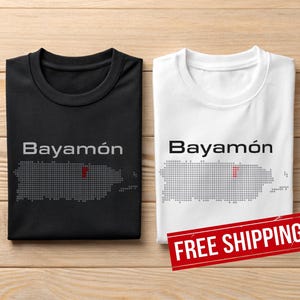 May include: Two t-shirts, one black and one white, each displaying the word "Bayamón" above a grey dotted outline of Puerto Rico with a red dot. The shirts are on a wooden surface.