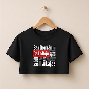 Könnte beinhalten: Schwarzes Crop-Top mit dem Text "SanGermán Cabo Rojo Lajas" in Weiß und Rot. Zusätzlicher Text in Weiß enthält "Cabor, Moca, Adj, Hormig, Coam, Guánica, Maricao, Peñuelas, aGrande, Yagüez". Das Shirt hängt an einem Holzkleiderbügel.