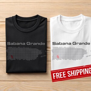 May include: Two t-shirts, one black and one white, featuring the text "Sabana Grande" above a pixelated map of Puerto Rico. The black shirt has a grey map and text, while the white shirt has a grey map and text with a red accent.