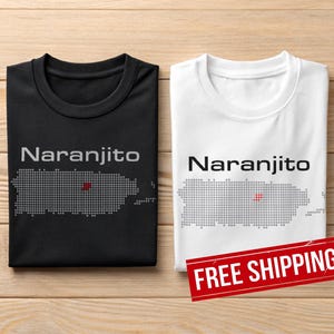 Könnte beinhalten: Zwei T-Shirts, eines schwarz und eines weiß, jeweils mit dem Wort "Naranjito" über einer gepunkteten Umrisslinie von Puerto Rico. Ein roter Punkt markiert einen Ort auf der Karte. Die Shirts liegen auf einer Holzoberfläche.