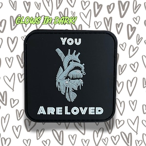 May include: Black patch with a white anatomical heart and the text "YOU ARE LOVED" in white. The patch glows in the dark.