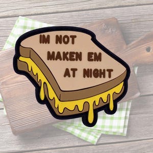 Peut inclure: Un écusson fantaisie en forme de sandwich au fromage grillé avec le texte "IM NOT MAKEN EM AT NIGHT". Le sandwich a du pain brun, du fromage jaune et un bord noir. L'écusson est posé sur une planche à découper en bois.