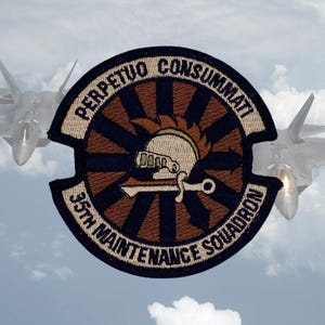 May include: A tan and brown embroidered patch with a dark blue circle in the center. The circle has a sunburst design with 16 rays. A helmet with a plume is in the center of the circle. A sword is behind the helmet. The text "PERPETUO CONSUMMATI" is above the circle and "35TH MAINTENANCE SQUADRON" is below the circle.