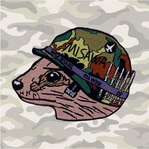 Könnte beinhalten: Ein braunes und rosa Cartoon-Wiesel, das einen grünen, braunen und gelben Tarnhelm mit dem Text "Misawa" und "Wolf Weasel" trägt. Der Helm hat ein violettes Band mit dem Text "YSA WOOW" und eine kleine Flugzeuggrafik auf der Seite. Das Wiesel trägt ein violettes Band mit dem Text "GP" und eine Reihe von Kugeln auf der Seite.