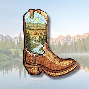 May include: An embroidered patch shaped like a brown cowboy boot. The shaft of the boot features a detailed landscape scene with a winding blue river, green pine trees, rolling hills, and a yellow sun set against a light blue sky background.