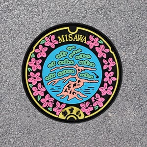 May include: A black and white manhole cover with a colorful design. The design features a stylized tree with pink flowers around the edge. The text "MISAW" is written in white letters on the cover.