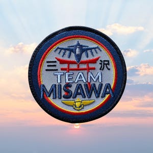 Könnte beinhalten: Runder, gestickter Aufnäher mit blauem Rand. Das Design umfasst ein rotes und weißes Torii-Tor, ein blaues Flugzeug und den Text "TEAM MISAWA". Der Aufnäher hat einen hellgrauen Hintergrund und ein gelbes Emblem.