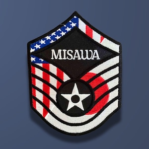May include: Embroidered patch with the word "MISAWA" in white on a black diamond. The patch features red and white stripes, a red circle, a white star, and a blue section with white stars, all on a dark blue background.