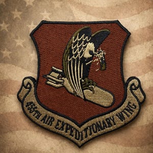 Op de afbeelding: Geborduurde patch met een adelaar die een bundel draagt, geplaatst op een schildvorm met een bruine en zwarte rand. De tekst "455th AIR EXPEDITIONARY WING" is aan de onderkant gebogen. De achtergrond toont een vervaagde Amerikaanse vlag.