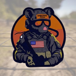 May include: A black and blue embroidered patch with a cartoon mouse wearing a military uniform and holding a rifle. The mouse is wearing a helmet with the words "Ready Airman Trained" on it. The patch is set against a sunset background.