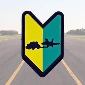 May include: A yellow and teal patch with a black border. The patch features a silhouette of a vehicle towing an airplane on the left side and a jet on the right side. The background shows an airport runway.