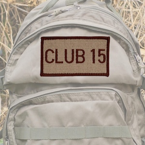 Könnte beinhalten: Ein rechteckiger Aufnäher mit dem Text "CLUB 15" in Dunkelbraun auf beigem Hintergrund gestickt. Der Aufnäher ist mit einer dunkelbraunen Umrandung versehen und an einem beigen Rucksack befestigt.