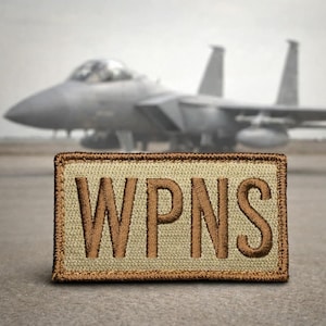 Pode incluir: Um patch bordado bege com a palavra "WPNS" em letras marrons. O patch é retangular com uma borda marrom. Um caça cinza está ao fundo.