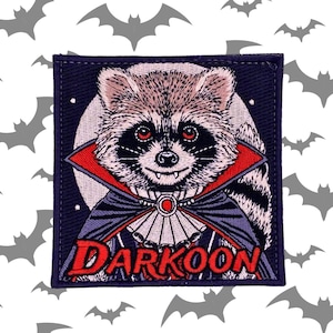 Peut inclure: Un patch brodé noir et bleu représentant un raton laveur déguisé en vampire avec une cape rouge et le texte "DARKOON".