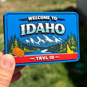 Può includere: Una toppa rettangolare blu con le parole "WELCOME TO IDAHO" sopra una scena di montagna e fiume. La toppa presenta anche una patata e il testo "TRVL ID".
