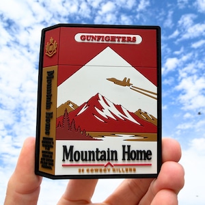 Puede incluir: Un parche rectangular de PVC con un diseño en rojo, blanco y bronceado que muestra un paisaje montañoso, un avión de combate y el texto "GUNFIGHTERS", "Mountain Home" y "20 COWBOY KILLERS". El parche tiene un borde negro y un emblema dorado.