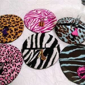 May include: Six round coasters with different animal print designs. Each coaster has a small charm attached, including a high heel shoe, a purse, and a dress.