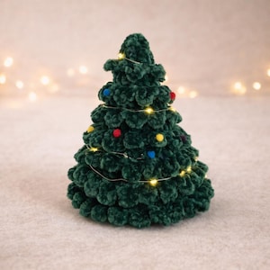 Könnte beinhalten: Ein handgefertigter, dunkelgrüner gehäkelter Weihnachtsbaum mit bunten Pompons und Lichterketten. Der Baum ist eine festliche Dekoration, perfekt für die Weihnachtszeit. Der Baum ist etwa 15 cm hoch.