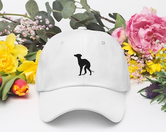 Op de afbeelding: Witte baseballcap met een zwart silhouet van een hond. De pet wordt omringd door kleurrijke bloemen, waaronder gele narcissen, roze pioenrozen en groen. Een casual accessoire.