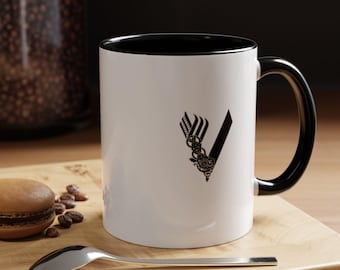 May include: White ceramic mug with a black rim and handle. The mug features a black and white graphic of the "Vikings" logo, a stylized "V" with a knot design.