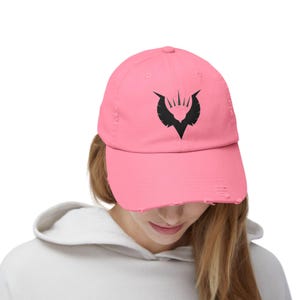 May include: A pink distressed baseball cap with a black embroidered design of a stylized bird with a crown on its head.