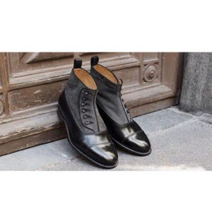 Può includere: Un paio di stivaletti in pelle nera con un pannello in camoscio grigio e dettagli di bottoni. Gli stivaletti hanno un design classico con punta arrotondata e tacco piccolo. Gli stivaletti sono esposti su una superficie grigia.