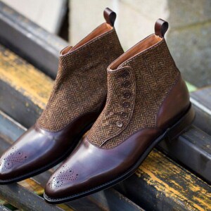 Può includere: Stivaletti in pelle marrone con pannelli in tweed. Gli stivali presentano una chiusura con bottoni, linguetta e dettagli brogue sulla punta. Gli stivali sono esposti su una superficie metallica. Design classico.