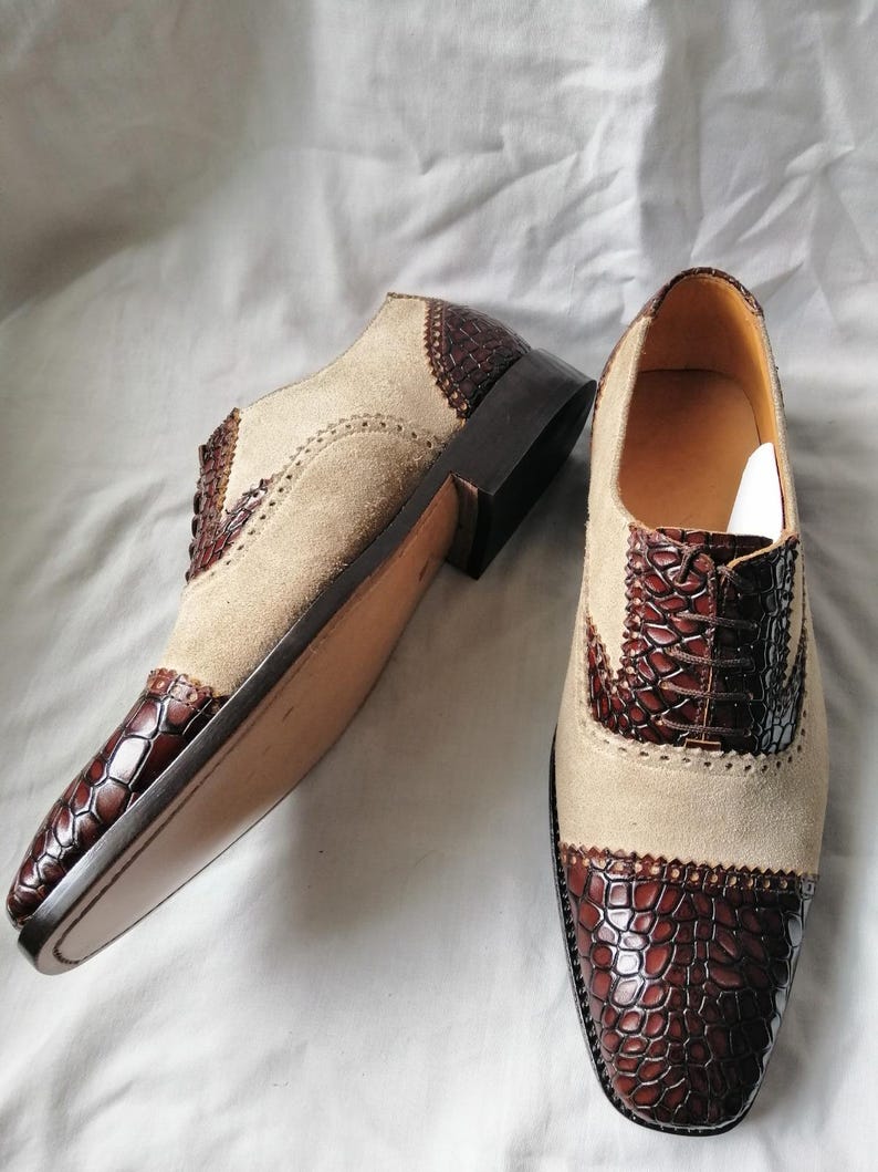 May include: A pair of brown and beige leather shoes with a crocodile pattern on the toe and heel. The shoes have a lace-up closure and a brogue design.