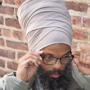 Puede incluir: Un hombre con un turbante gris, gafas de montura negra y una chaqueta gris. Tiene barba gris y se está ajustando las gafas con la mano derecha.