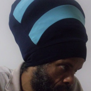 Puede incluir: Un hombre con un sombrero de color azul oscuro y azul claro con un patrón. El sombrero tiene una parte superior redondeada y un ala ancha. El hombre tiene barba y mira hacia un lado.