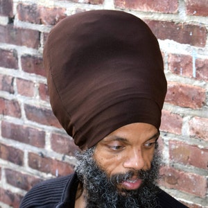 May include: A man wearing a brown fabric head wrap, also known as a durag, with a long gray beard. He is wearing a black shirt.