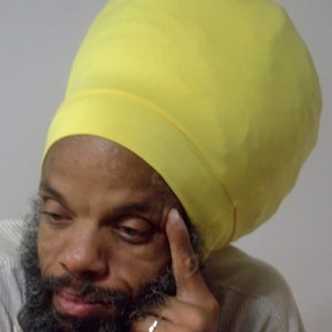 Puede incluir: Un hombre con un turbante de color amarillo brillante. Tiene barba y mira hacia abajo.