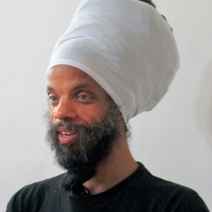 May include: A man with a long beard and a white turban is smiling. He is wearing a black shirt.
