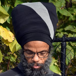 Puede incluir: Gorro negro con un diseño diagonal de pata de gallo blanco y negro. El gorro está hecho de un material suave. El fondo presenta hojas verdes y amarillas. El gorro lo lleva una persona con barba y gafas.