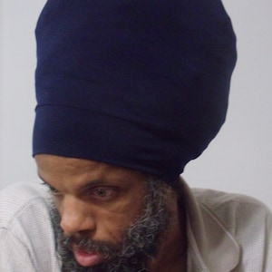 Puede incluir: Un hombre con una larga barba gris lleva un turbante azul oscuro y una camisa gris clara.