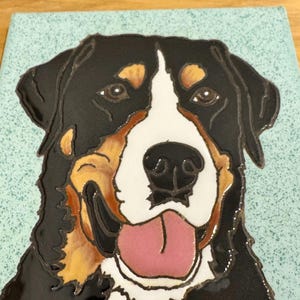 Puede incluir: Azulejo de cerámica con un retrato detallado de un Boyero de Berna. El perro tiene pelaje negro, blanco y marrón, con una lengua rosa. El azulejo tiene un fondo azul claro y es probablemente un artículo decorativo.