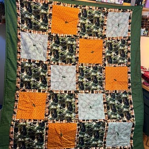 May include: A handmade quilt with a green border. The quilt features a patchwork design with squares of orange, gray, and a forest-themed fabric with bears. Each square has a decorative element. The quilt is displayed on a wall.