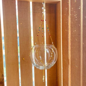 May include: A clear glass hanging planter with a gold chain and colorful beads. The planter is hanging from a hook in a wooden crate.