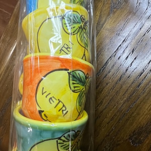 May include: A stack of five ceramic cups wrapped in clear plastic. Each cup features a hand-painted yellow lemon design with green leaves and the text VIETRI. The cups have different colored rims and bases, including blue, yellow, orange, and teal.
