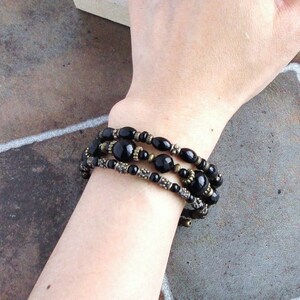 May include: A layered bracelet with black beads and gold accents. The bracelet is made of multiple strands of beads, with some beads being larger than others. The bracelet is worn on a person's wrist.
