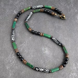 Op de afbeelding: Een kralenketting met cilindrische zwarte kralen met witte geëtste bladpatronen, afgewisseld met rechthoekige groene stenen kralen, kleine ronde zwarte kralen en getextureerde bruine cilindrische tussenstukjes, afgewerkt met een messing karabijnsluiting.