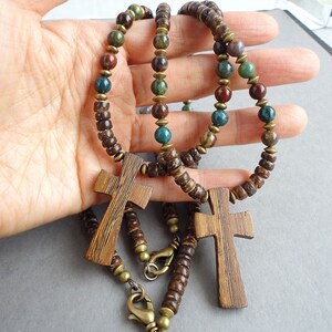 May include: Two wooden cross pendants on brown and green beaded necklaces. The crosses are made of dark wood and have a grain pattern. The beads are round and have a natural, earthy look.