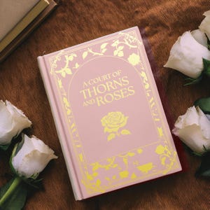 May include: A pink hardback book with gold lettering that reads "A Court of Thorns and Roses". The book is surrounded by white roses.