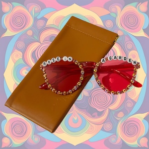 Puede incluir: Gafas de sol redondas rojas con pedrería y las palabras "ROSE IN A GARDEN" escritas en las monturas.