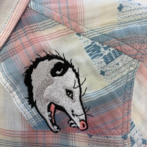 May include: Close-up of a plaid shirt in pastel pink, blue, and white. The shirt features an embroidered opossum patch on the pocket. The opossum is gray and white with a pink mouth and black accents.